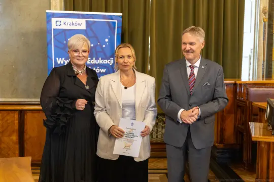 Trzy osoby stojące razem, jedna z dyplomem, w tle baner z logo Krakowa.