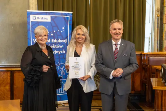 Trzy osoby trzymają dyplom w sali z zielonymi zasłonami i banerem Krakowa w tle.