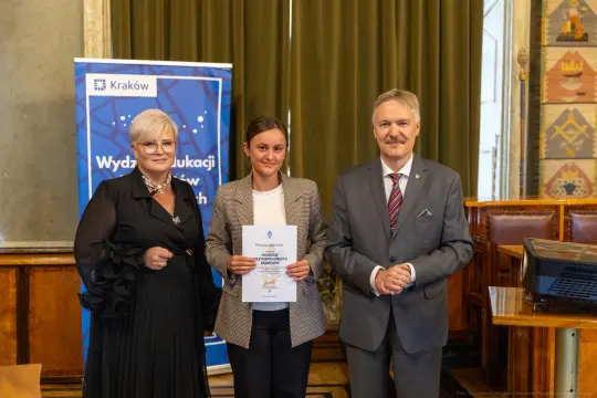 Uroczystość wręczenia dyplomu w Krakowie, trzy osoby pozujące z dokumentem.