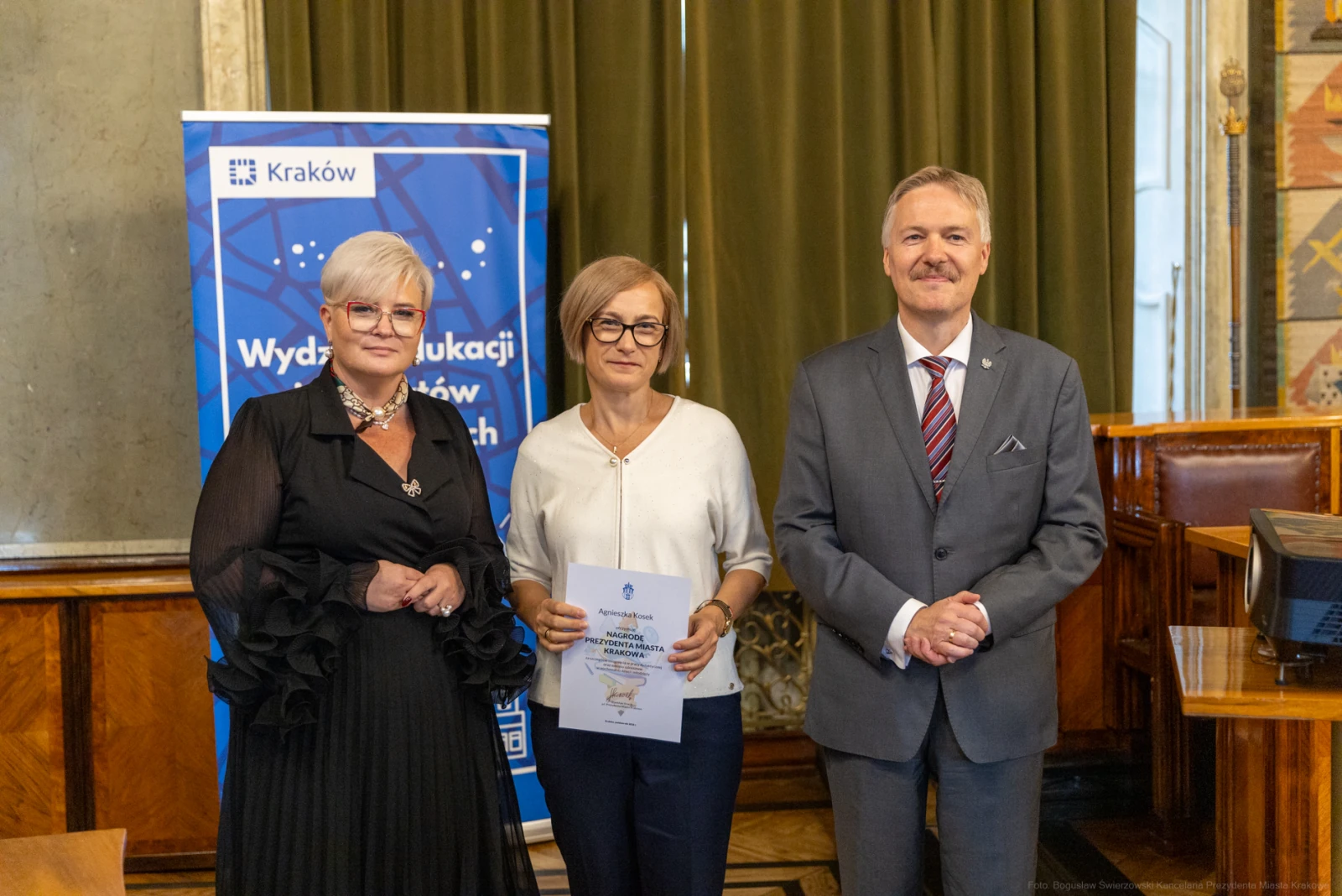 Trzy osoby w formalnych strojach, trzymające dokument, w tle baner z logo Krakowa.  Autor: Bogusław Świerzowski