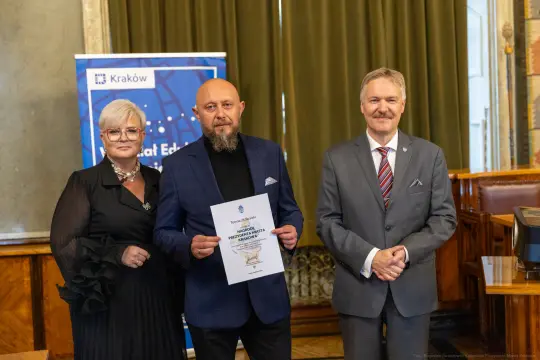 Trzy osoby w formalnych strojach trzymają dokument w sali z zielonymi zasłonami.