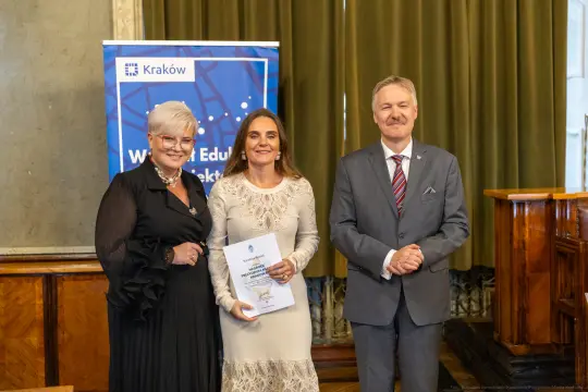 Trzy osoby w formalnych strojach, trzymające dokumenty, w eleganckim wnętrzu.