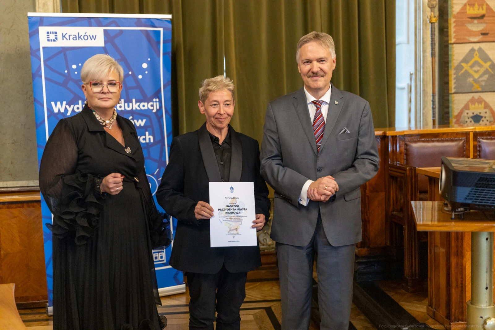 Trzy osoby w formalnych strojach trzymają dyplom w sali z dekoracjami.  Autor: Bogusław Świerzowski