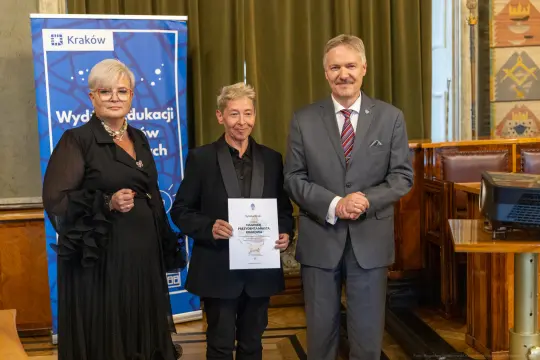 Trzy osoby w formalnych strojach trzymają dyplom w sali z dekoracjami.