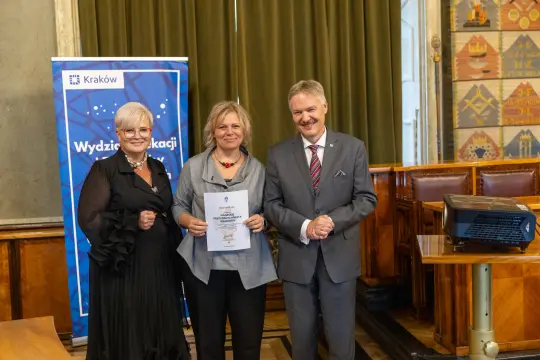 Trzy osoby w formalnych strojach, trzymające dyplom w sali z dekoracjami.