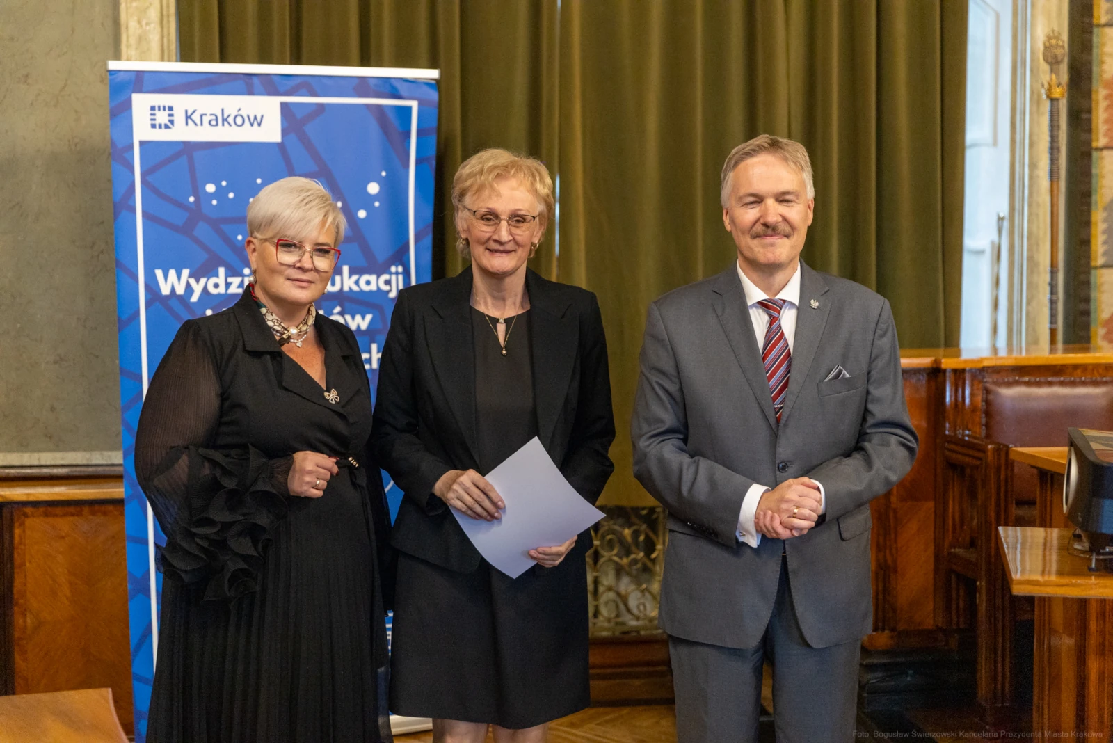 Trzy osoby w formalnych strojach, stojące obok siebie, z uśmiechami, w tle baner z logo Krakowa.  Autor: Bogusław Świerzowski