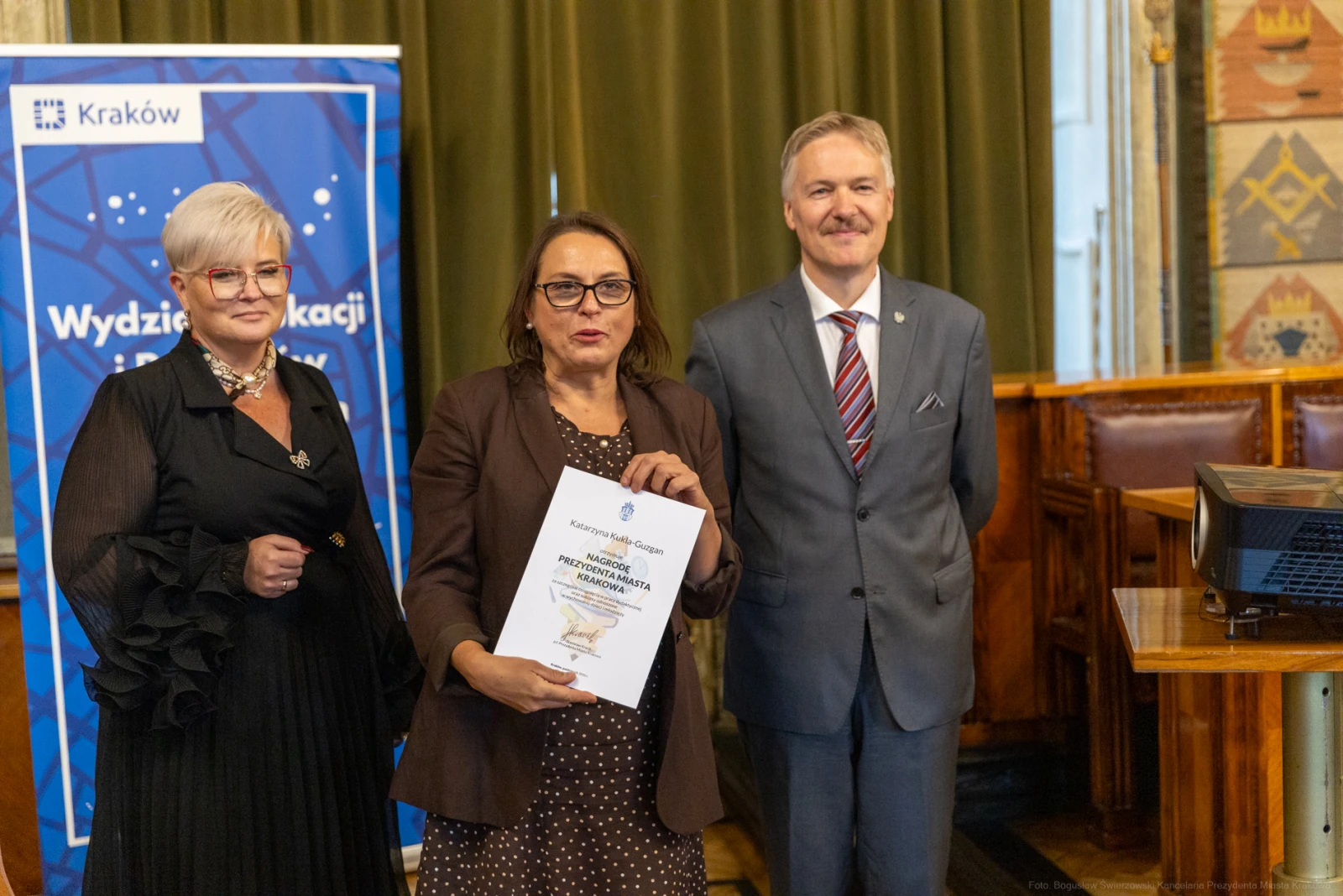 Trzy osoby trzymają dokument w sali z dekoracjami, obok baner z logo Krakowa.  Autor: Bogusław Świerzowski