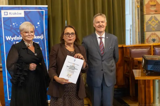 Trzy osoby trzymają dokument w sali z dekoracjami, obok baner z logo Krakowa.