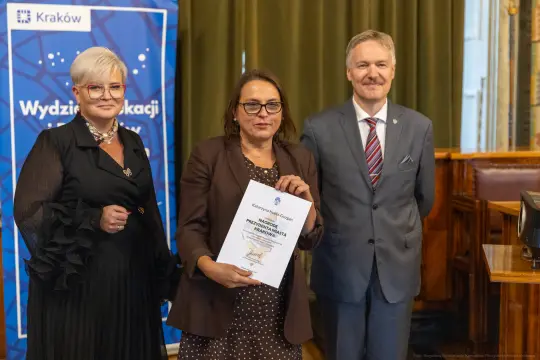 Trzy osoby w formalnych strojach, jedna trzyma dyplom, w tle baner z logo Krakowa.