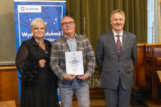Trzy osoby w formalnych strojach, trzymające dyplom w sali z zielonymi zasłonami.