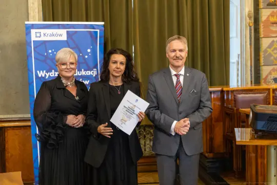 Trzy osoby w formalnych strojach, trzymające dokumenty, w tle banner z logo Krakowa.