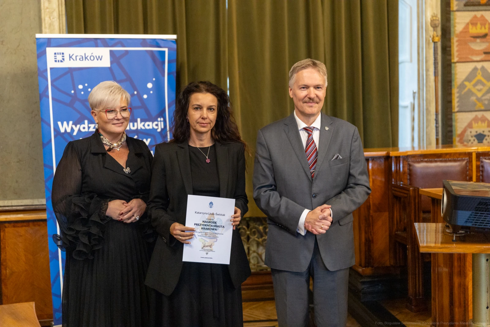Trzy osoby w formalnych strojach, trzymające dyplom w sali z dekoracjami.  Autor: Bogusław Świerzowski