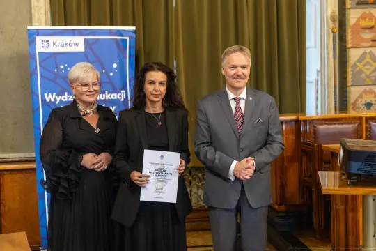 Trzy osoby w formalnych strojach, trzymające dyplom w sali z dekoracjami.