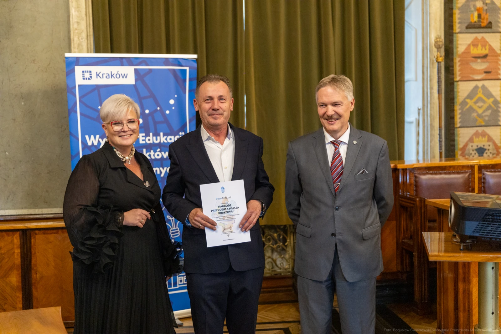 Trzy osoby w formalnych strojach trzymają dyplom w sali z dekoracjami.  Autor: Bogusław Świerzowski