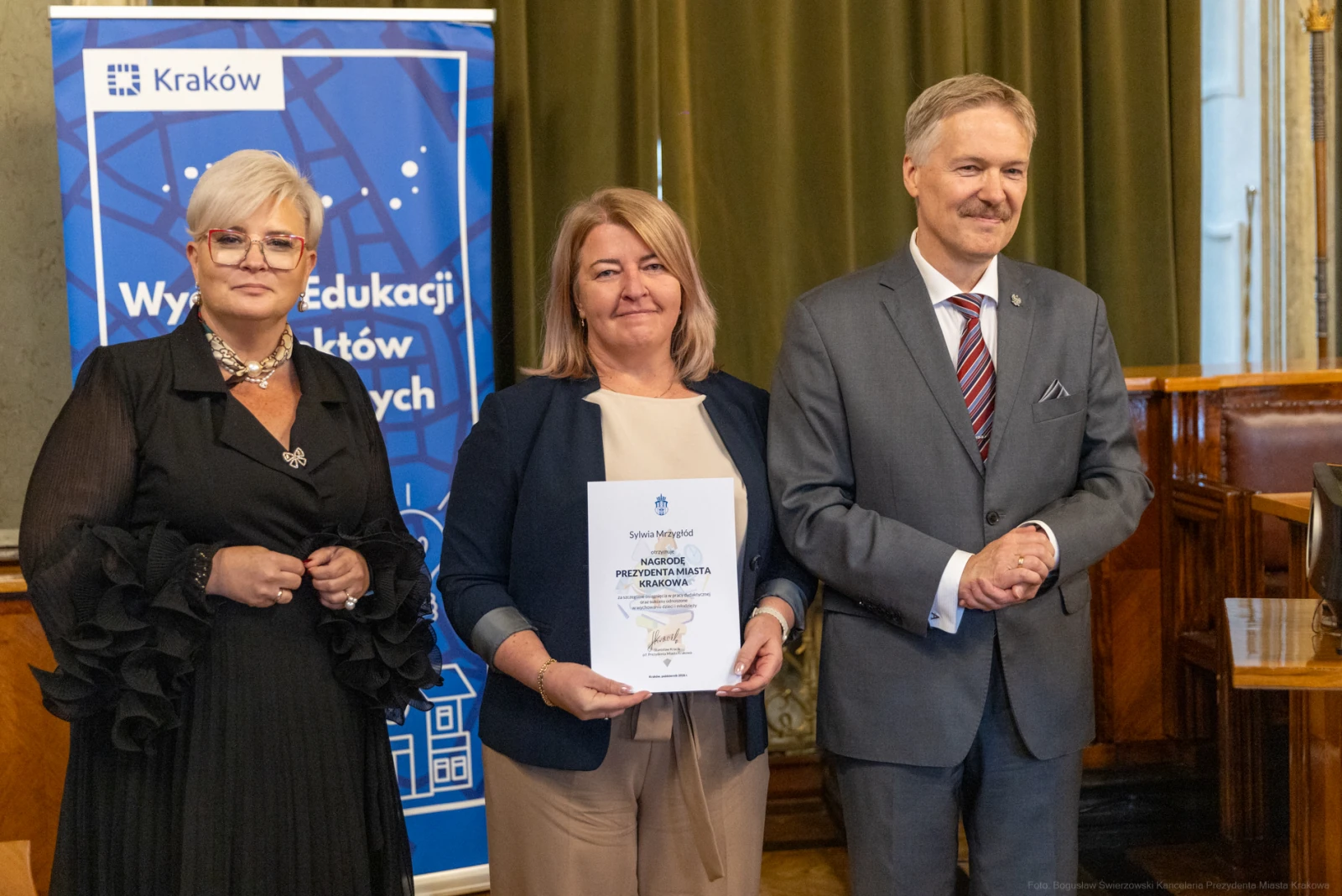 Trzy osoby w formalnych strojach, jedna z dyplomem, w tle baner z logo Krakowa.  Autor: Bogusław Świerzowski