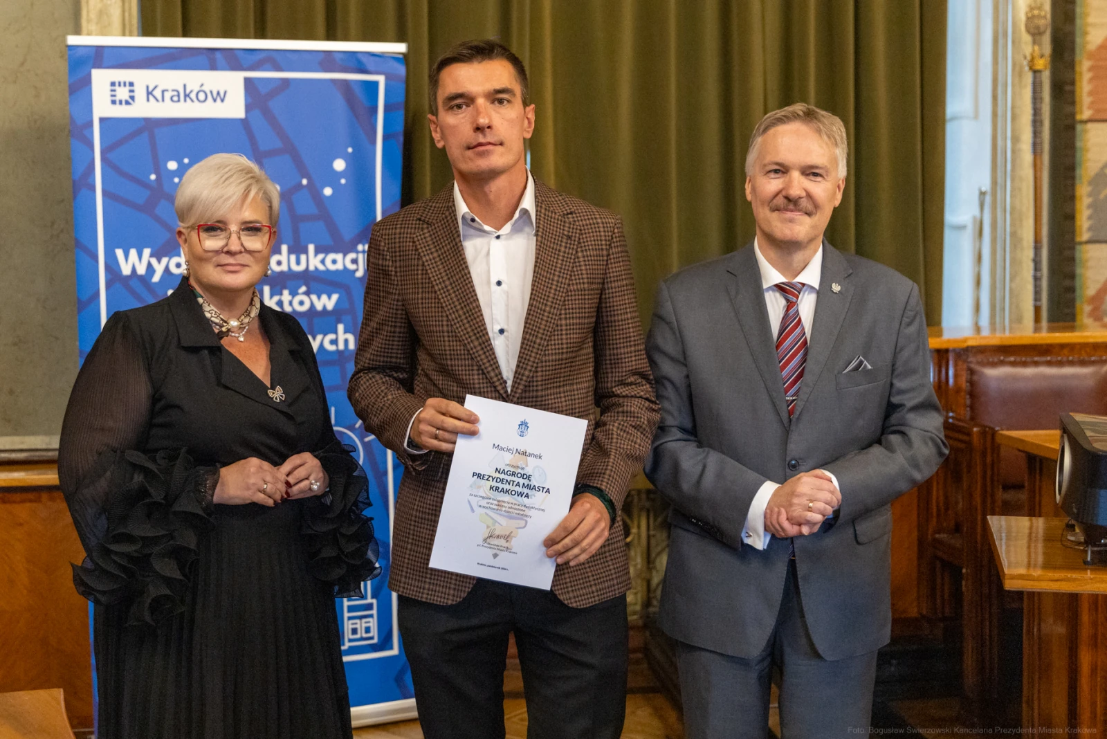 Mężczyzna z dyplomem, obok dwie osoby, w tle dekoracje sali.  Autor: Bogusław Świerzowski