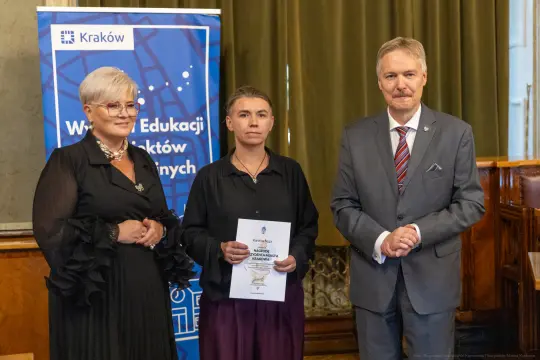 Wręczenie nagrody w Krakowie, trzy osoby z dokumentem i banerem edukacyjnym w tle.