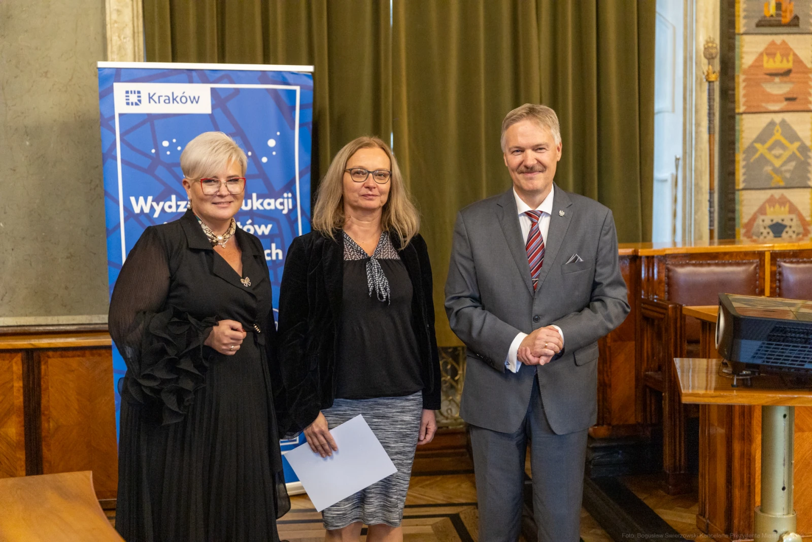 Trzy osoby w formalnych strojach stoją obok siebie w sali z dekoracjami, w tle baner Wydziału Edukacji.  Autor: Bogusław Świerzowski