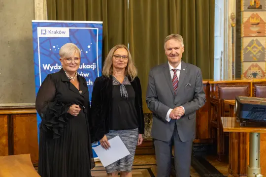 Trzy osoby w formalnych strojach stoją obok siebie w sali z dekoracjami, w tle baner Wydziału Edukacji.
