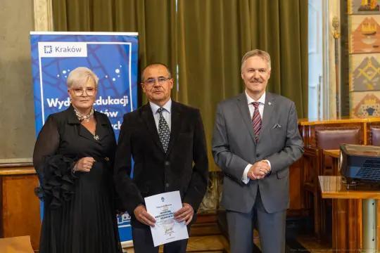 Trzy osoby w formalnych strojach, jedna z dyplomem, w tle baner z logo Krakowa.