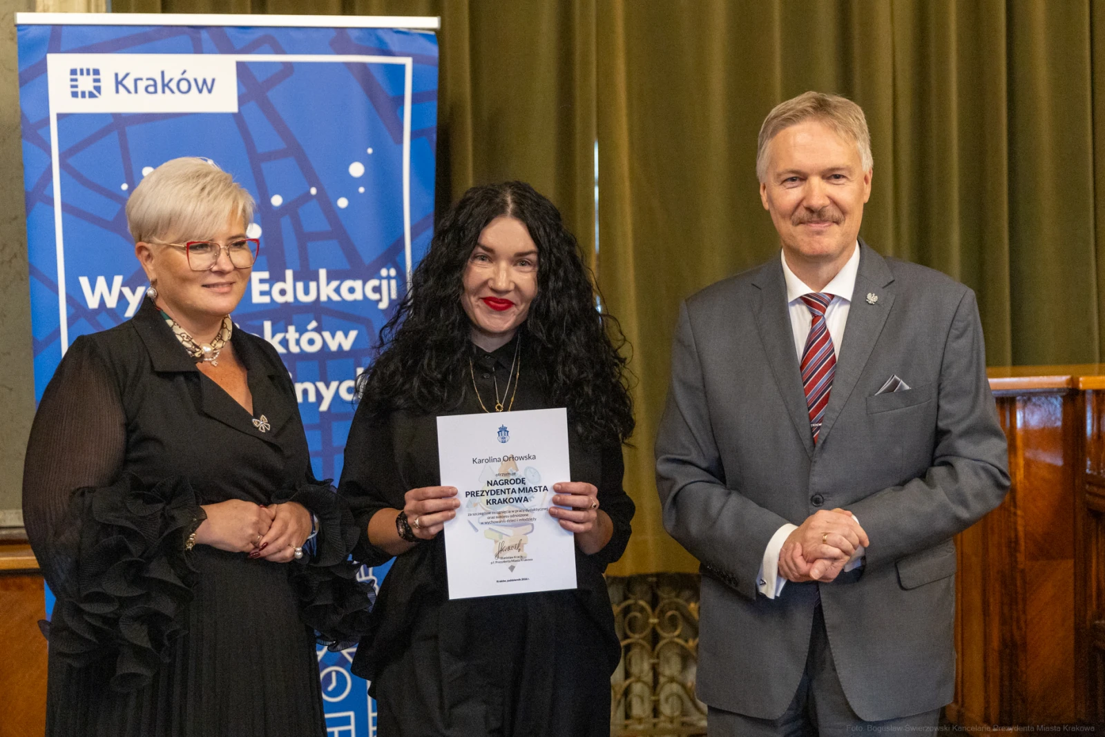 Kobieta z certyfikatem, obok niej dwie osoby w formalnych strojach, tło z logo Krakowa.  Autor: Bogusław Świerzowski