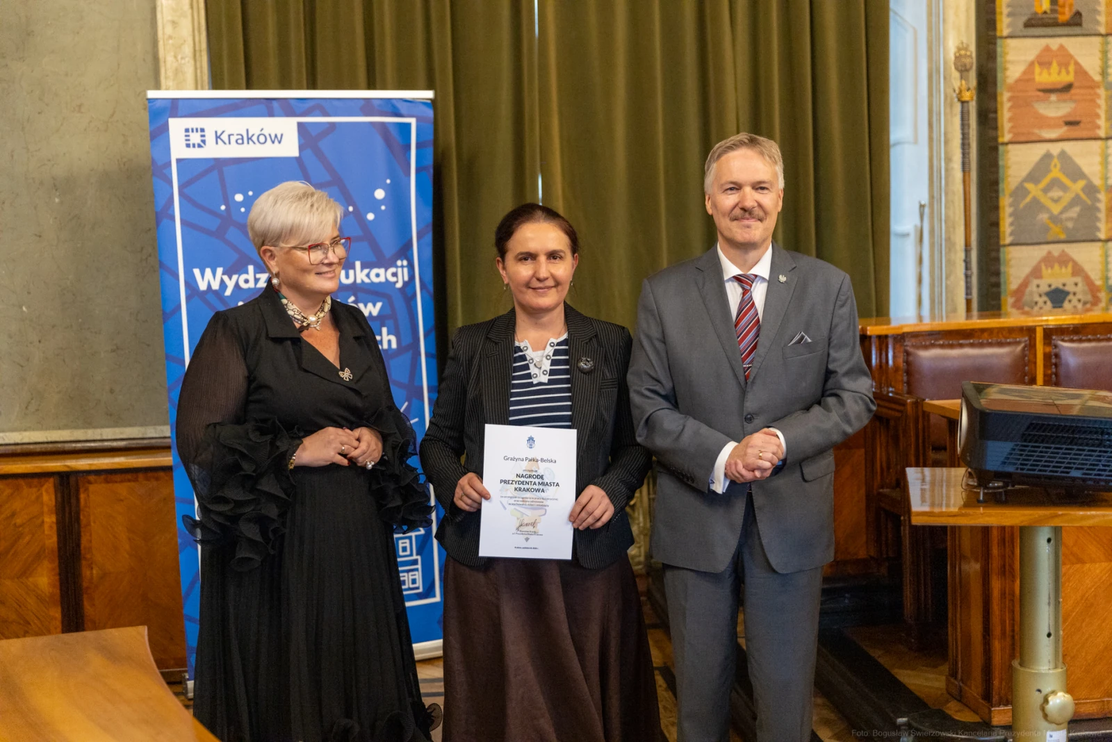 Trzy osoby w formalnych strojach, trzymające dyplom w sali z dekoracjami.  Autor: Bogusław Świerzowski