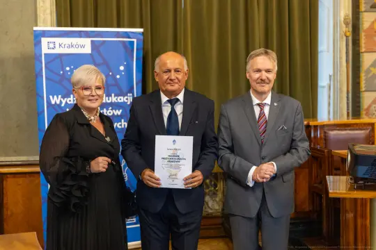 Trzy osoby stojące obok siebie, trzymające dyplom w sali z zielonymi zasłonami.