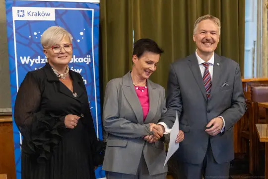 Trzy osoby w formalnych strojach, uśmiechające się i trzymające dokumenty. Tło z logo Krakowa.