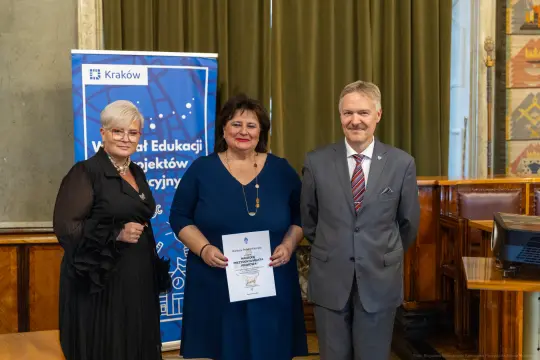 Trzy osoby w formalnych strojach, jedna z dyplomem, w tle banner i zasłony.