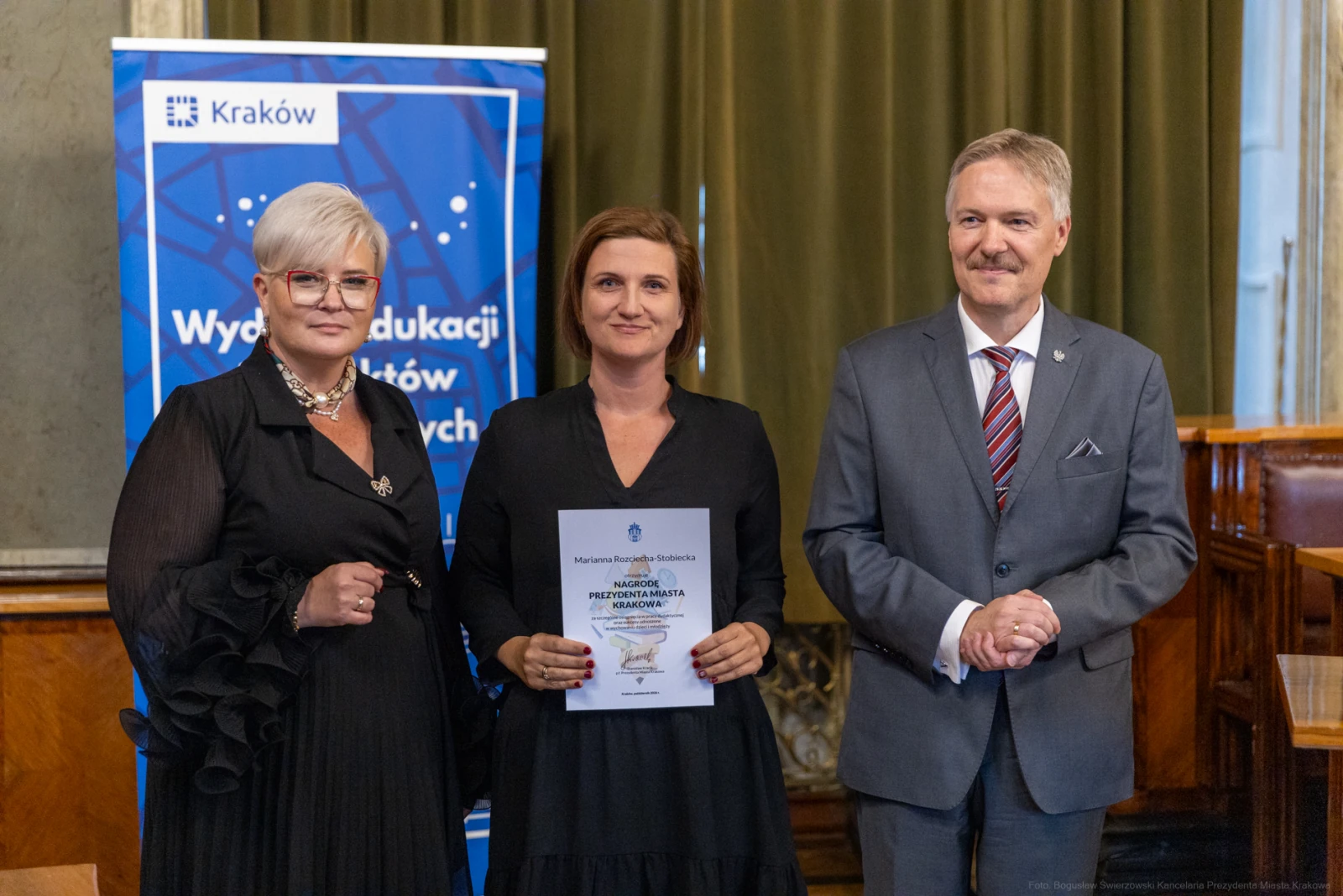 Wręczenie nagrody w Krakowie, trzy osoby z dyplomem i banerem w tle.  Autor: Bogusław Świerzowski