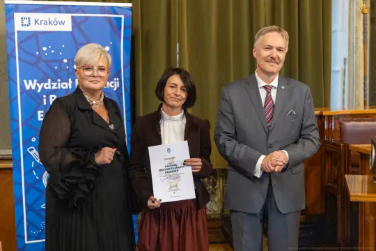 Trzy osoby trzymają dyplom w sali z zielonymi zasłonami. W tle baner Wydziału Edukacji.