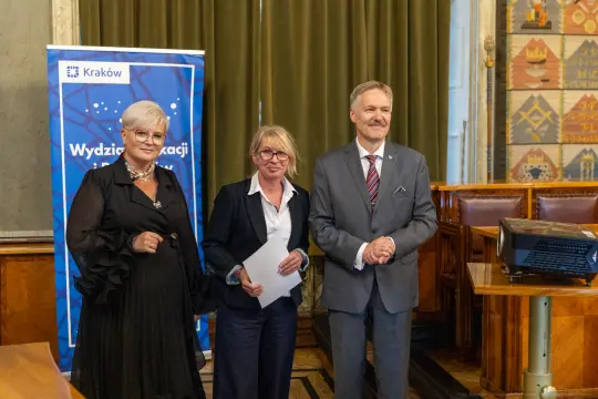 Trzy osoby w formalnych strojach stoją obok siebie w sali z dekoracjami.