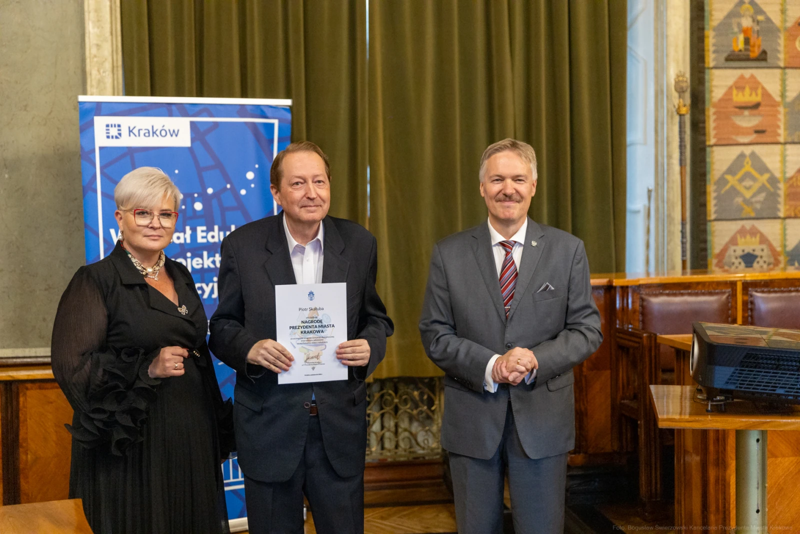 Trzech mężczyzn i kobiet w formalnych strojach, trzymających dokumenty w sali z dekoracjami.  Autor: Bogusław Świerzowski
