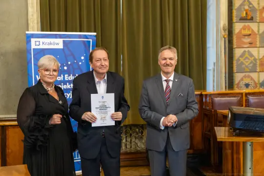 Trzech mężczyzn i kobiet w formalnych strojach, trzymających dokumenty w sali z dekoracjami.