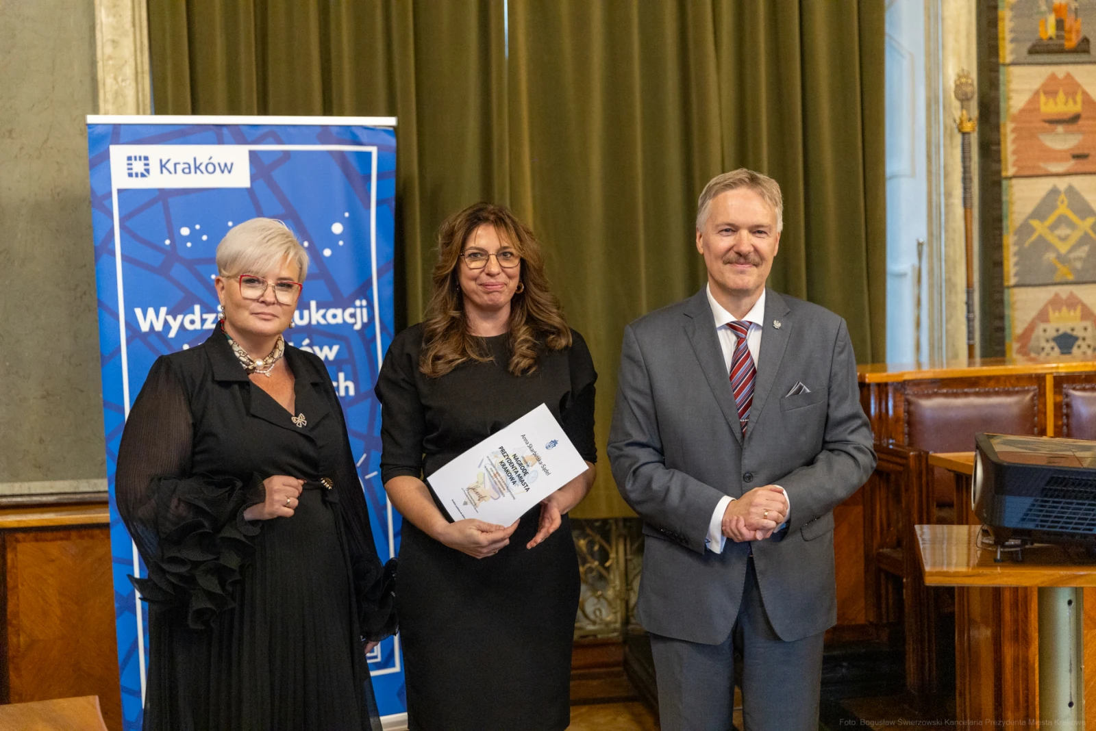 Trzy osoby w formalnych strojach stoją obok siebie, trzymając dokumenty, w tle banner i eleganckie wnętrze.  Autor: Bogusław Świerzowski