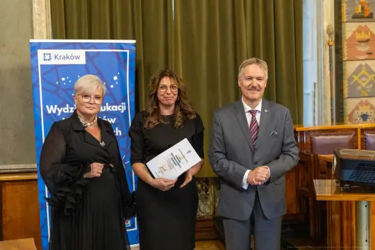 Trzy osoby w formalnych strojach stoją obok siebie, trzymając dokumenty, w tle banner i eleganckie wnętrze.