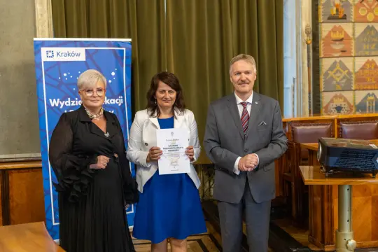 Trzy osoby w formalnych strojach, jedna trzyma dyplom, w tle dekoracyjne zasłony.