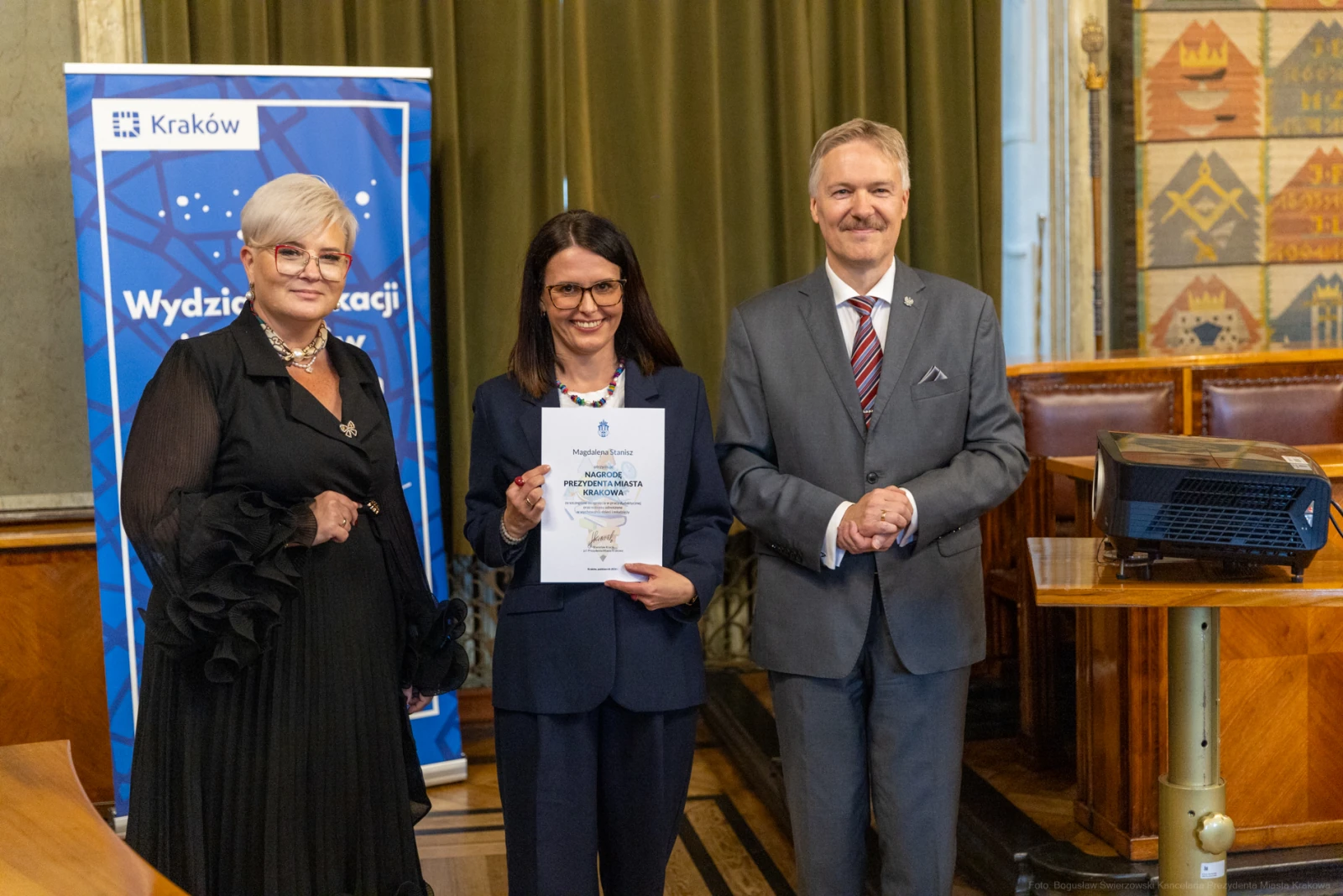 Trzy osoby w formalnych strojach trzymają dyplom w sali z dekoracjami.  Autor: Bogusław Świerzowski