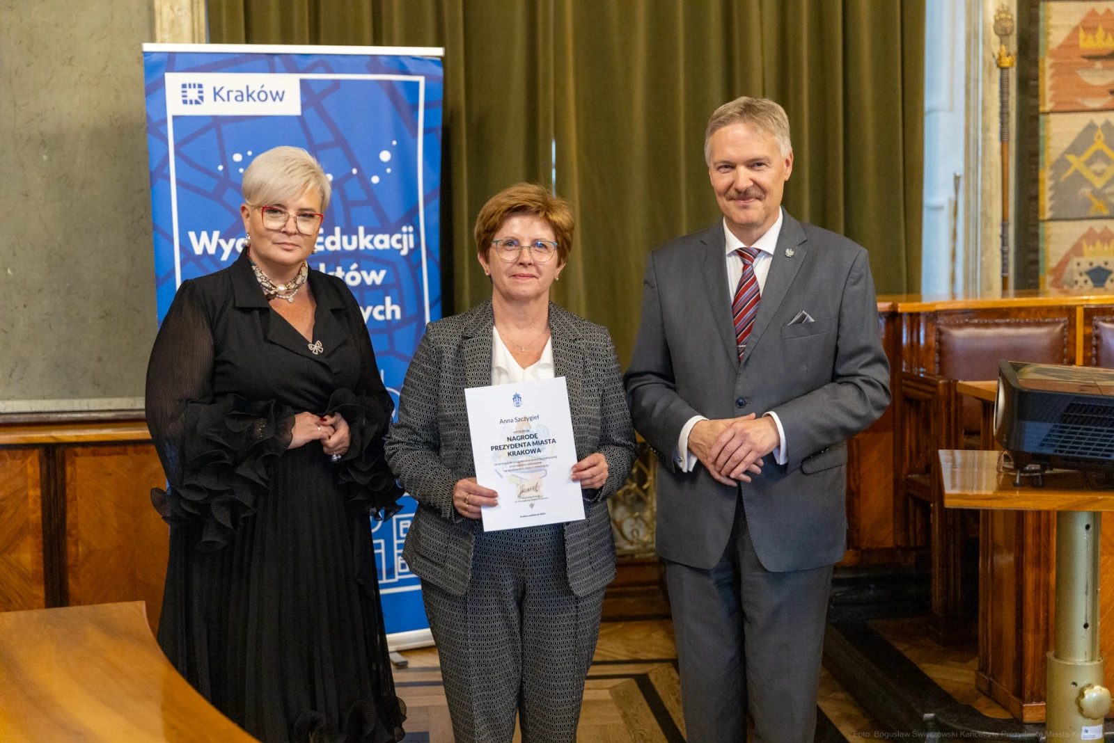 Trzy osoby w formalnych strojach, jedna z dyplomem, w tle flagi i herb Krakowa.  Autor: Bogusław Świerzowski