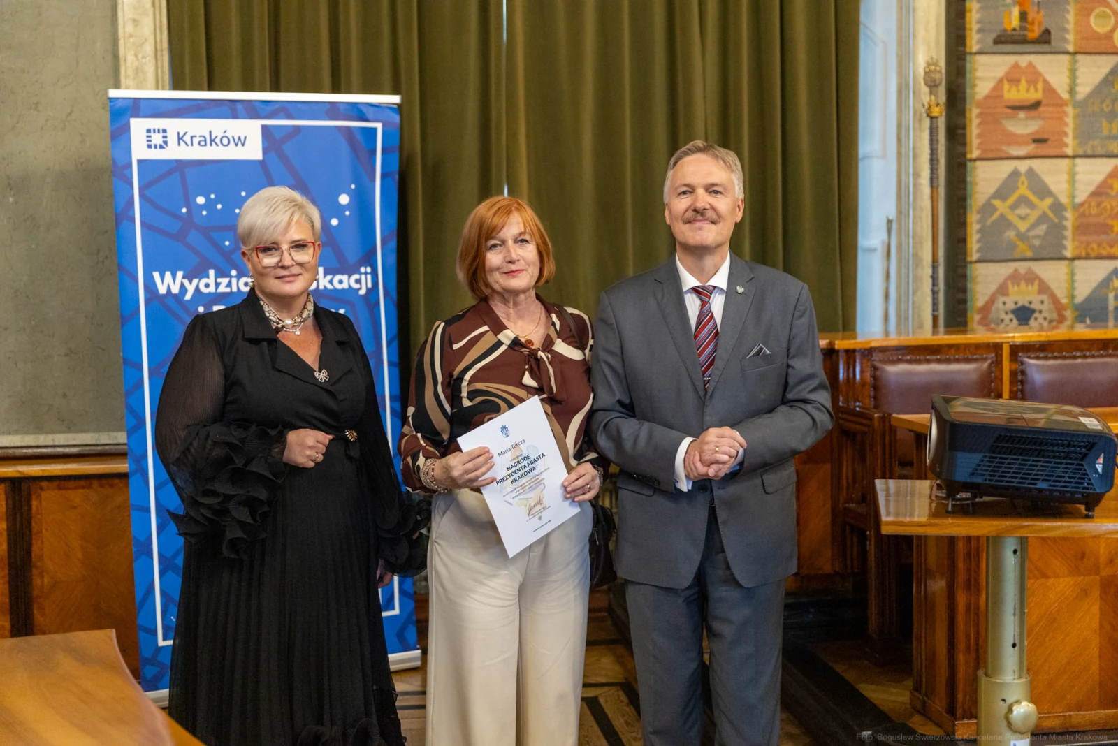 Trzy osoby w formalnych strojach, jedna z dyplomem, w tle baner Wydziału Edukacji Krakowa.  Autor: Bogusław Świerzowski