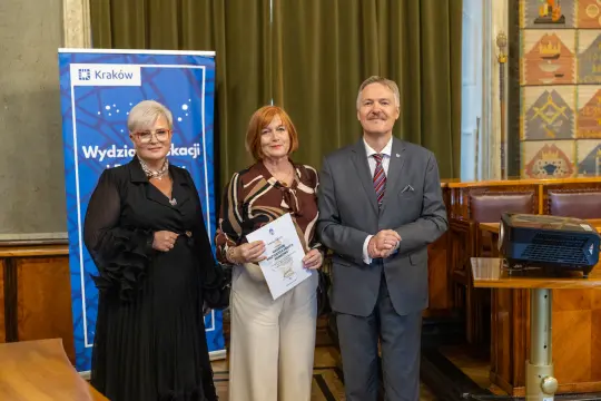 Trzy osoby w formalnych strojach, jedna z dyplomem, w tle baner Wydziału Edukacji Krakowa.