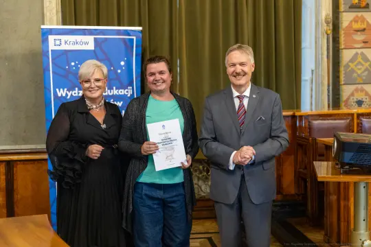 Wręczenie dyplomu w sali miejskiej, uśmiechnięte osoby w formalnych strojach.