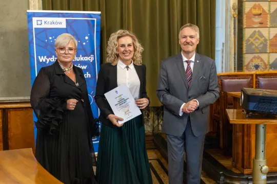 Trzy osoby w formalnych strojach trzymają dokument w sali z dekoracjami.
