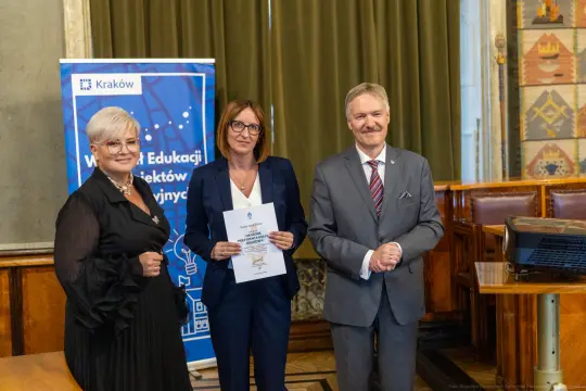 Trzy osoby w formalnych strojach, trzymające dokumenty, w tle baner z logo Krakowa.