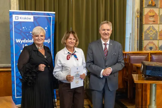 Wręczenie dyplomu w Krakowie, trzy osoby uśmiechają się, w tle baner Wydziału Edukacji.