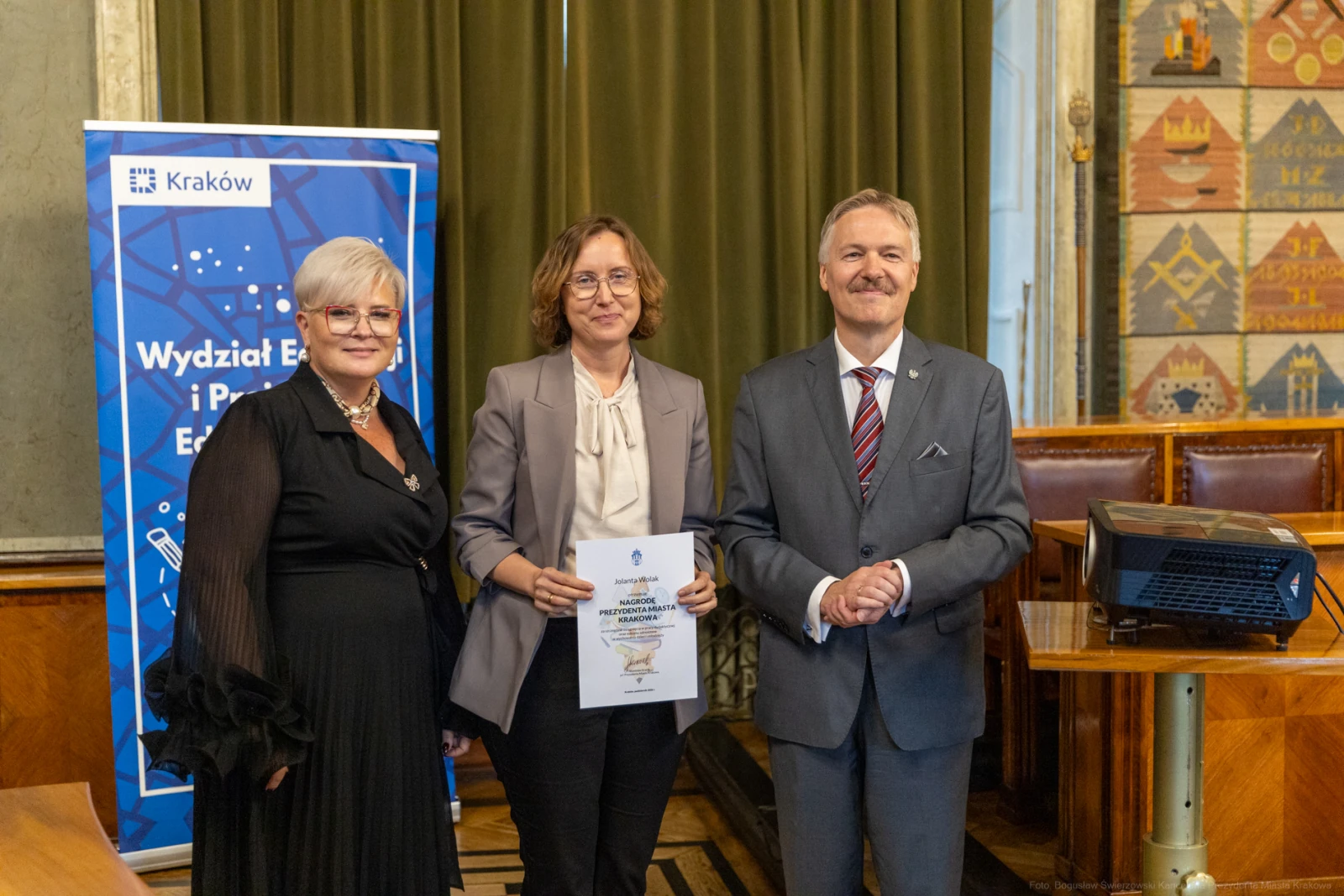 Trzy osoby w formalnych strojach, trzymające dokument, w tle baner i sala konferencyjna.  Autor: Bogusław Świerzowski