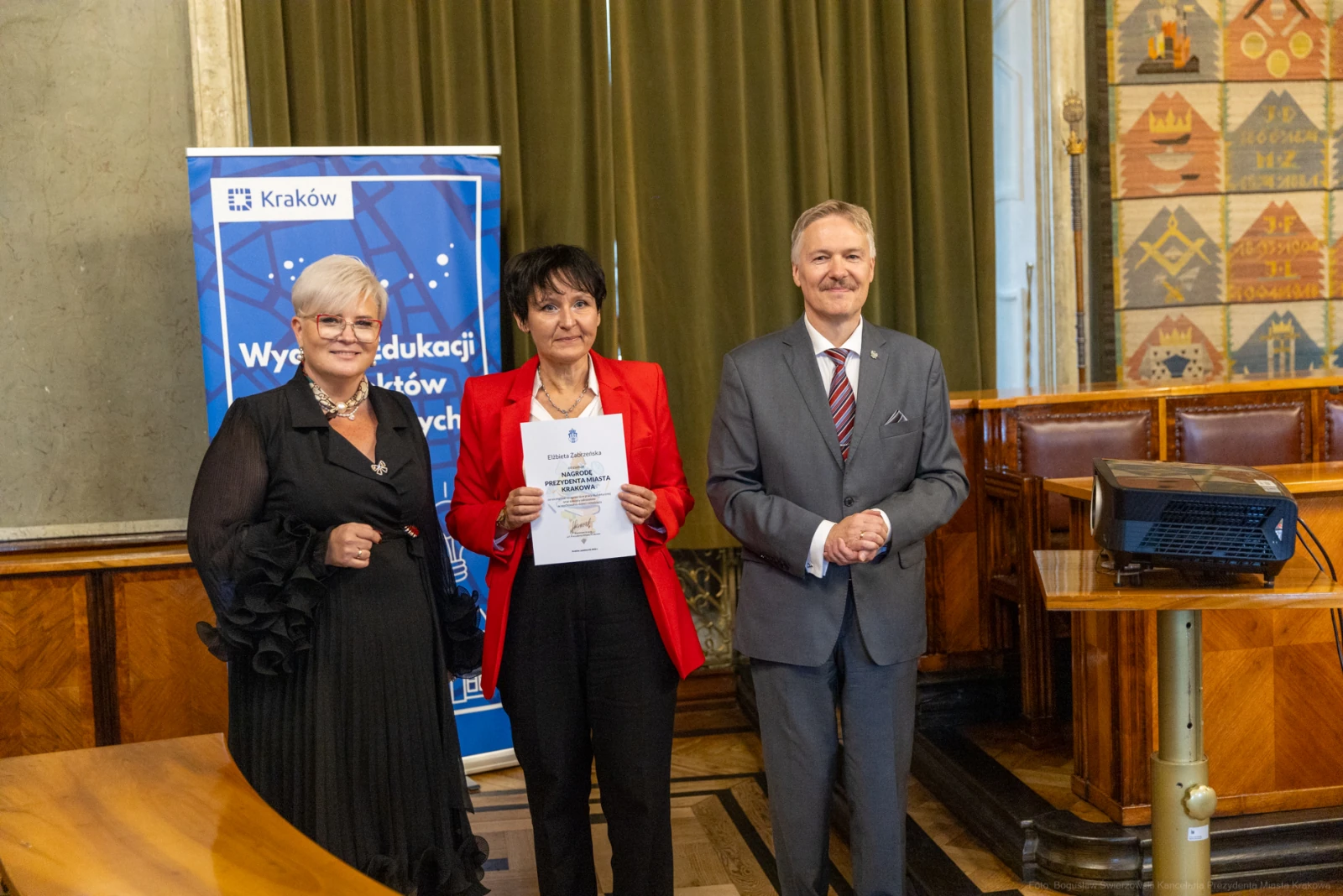 Trzy osoby w formalnych strojach trzymają dokumenty w sali z dekoracjami.  Autor: Bogusław Świerzowski