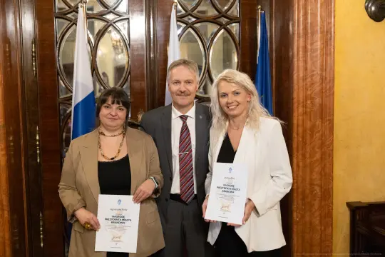 Trzy osoby trzymają dyplomy, stojąc w eleganckim wnętrzu z flagami w tle.