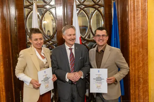 Trzy osoby z dyplomami, w tle flagi Polski i Unii Europejskiej.