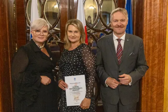 Trzy osoby w eleganckich strojach, trzymająca dokument w sali z dekoracyjnymi drzwiami.